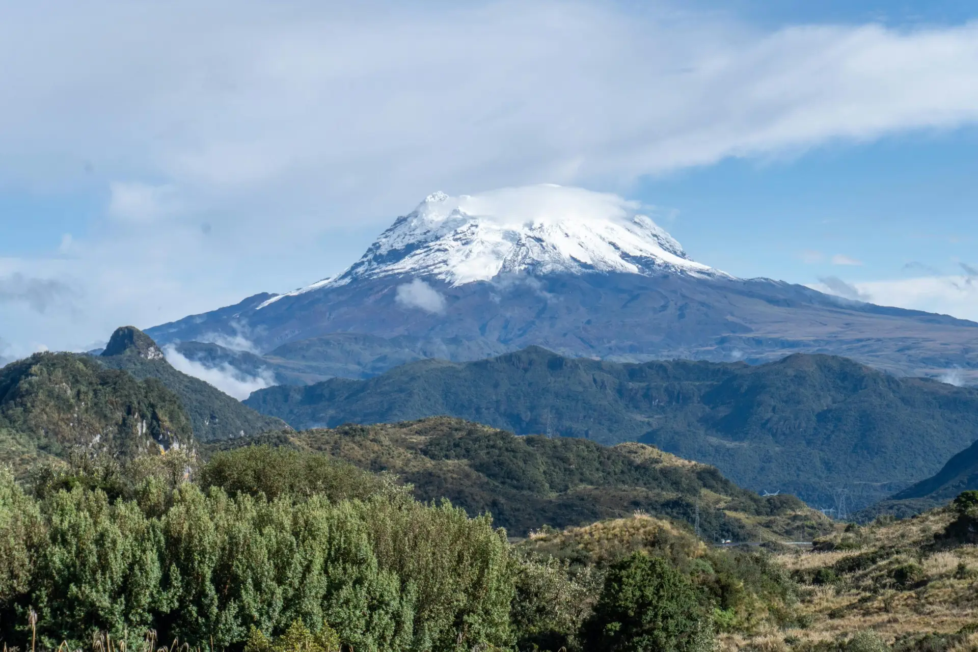 A breathtaking view of the snow-capped Cotopaxi Volcano emerging above lush green mountains under a clear sky in Ecuador.
